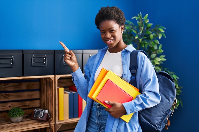 African american woman wearing student backpack and holding books smiling happy pointing with hand and finger to the side